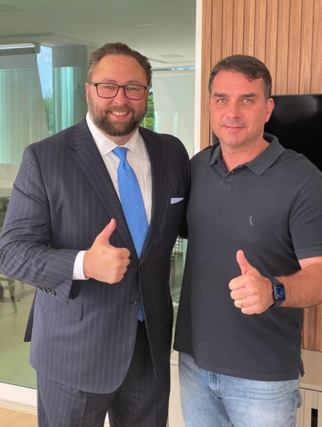 Imagem com Jason Miller, homem branco, barba, usando terno azul marinho e gravata azul fazendo sinal de positivo com a mão ao lado de FLávi Bolsonaro, homem branco, usando camisa polo cinza e calça jeans