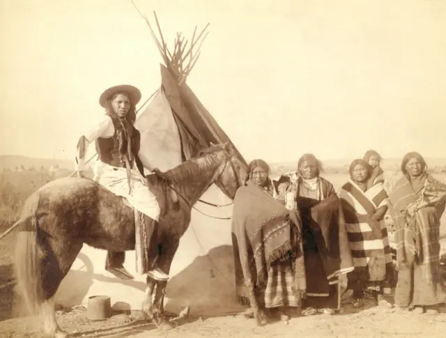 Ayakta duran dört Lakota kadını, beşiklerde bebek tutan üçü ve at sırtında bir Lakota adamının fotoğrafı. Muhtemelen 1891'de Pine Ridge Rezervasyonu'nda veya yakınında bir tipi'nin önündeler. Fotoğraf sepya rengindedir. 