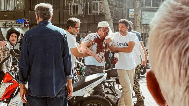 Dois homens ajudam um homem machucado, com a camisa suja de sangue e uma faixa na cabeça