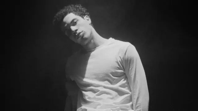 Imagen en blanco y negro de un hombre con una camiseta blanca de manga larga mirando a la cámara. Tiene el pelo corto, rizado y oscuro.