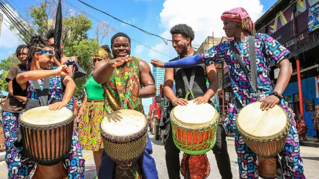 Quatre jeunes jouent du tambour. Ils sont debout en rang et vêtus de vêtements aux motifs colorés.