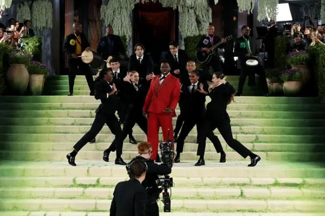 Grupo de personas baila y actúa en una escalinata verde durante un evento de gala; una persona en el centro viste traje rojo, rodeada de bailarines de negro, con músicos y cámaras al fondo.
