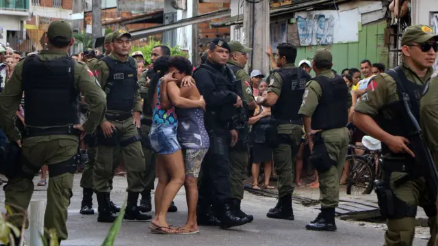 Duas mulheres abraçadasluto,meio a policiais, na periferiaBelém, após assassinato10 pessoas a tirosum bar