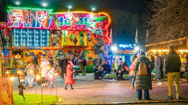 Atracciones y luces coloridas en el festival con adultos y niños caminando en primer plano.