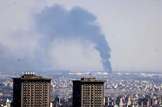 17 Haziran 2025'te İran'ın başkenti Tahran'a düzenlenen İsrail saldırısının ardından uzaktaki bir petrol rafinerisinden yükselen dumanlar ve yakın planda iki gökdelen
