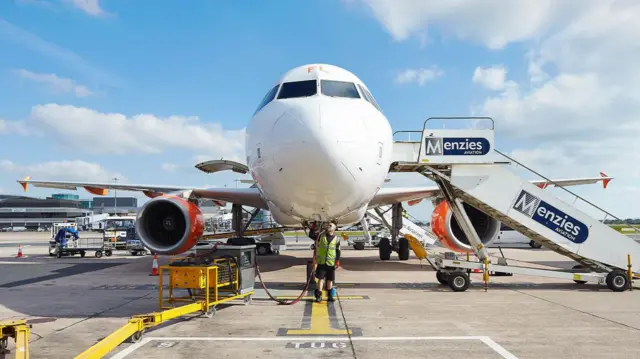 Vista frontal de um avião comercial estacionado, com equipes de solo trabalhando sob o nariz da aeronave e uma escada móvel com a marca “Menzies Aviation” posicionada ao lado