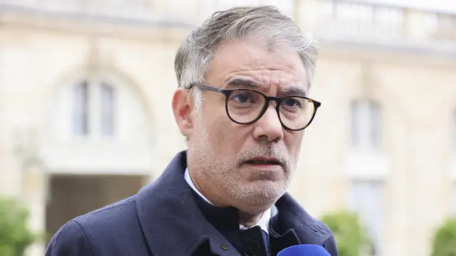 Оливье Фор в очках, с щетиной и в верхней одежде говорит в микрофон на фоне кремового здания