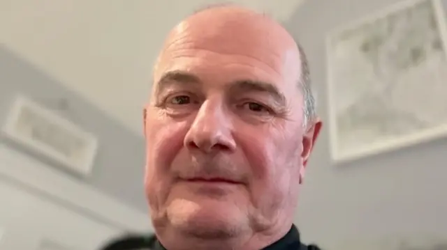 Un hombre calvo mira a la cámara. Detrás de él hay cuadros colgados en la pared.  