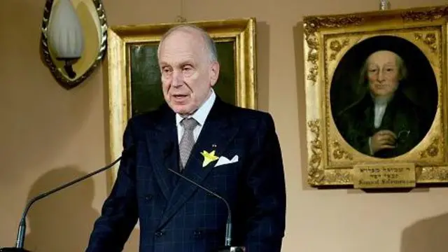 Рональд Лаудер стоїть за трибуною з мікрофонами, у чорному костюмі. За спиною у нього на бежевих стінах - кілька картин у рамках з позолотою та світильник.