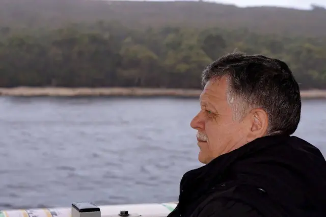 Чоловік середнього віку в темному одязі сидить на борту човна чи катера й дивиться у бік водної гладі. На задньому плані – смуга берега з густими деревами та пагорбами, оповитими легкою імлою.