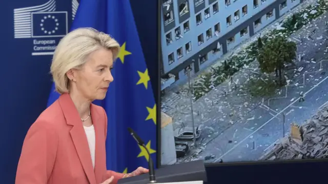 Урсула фон дер Ляйен в красном пиджаке на фоне флага ЕС и экрана, на котором видно здание с вылетевшими окнами в Киеве