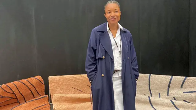 Kelechi Ejikeme, vêtu d'un long manteau bleu marine, se tient devant des tapis de jute - principalement bruns et beiges avec des motifs bordés de bleu marine - lors du salon Decorex à Londres. 