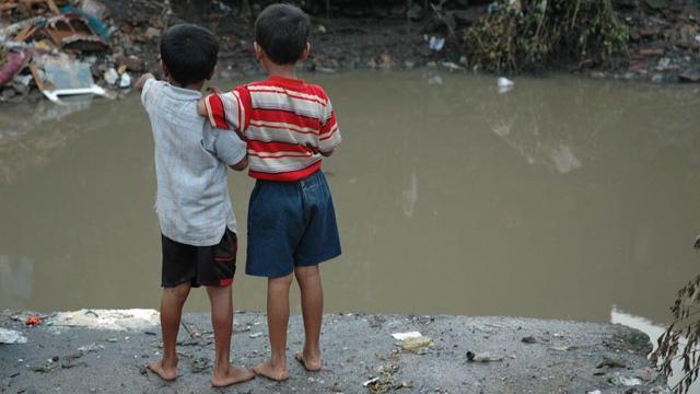 crianças descalças em frente à água parada e lixo