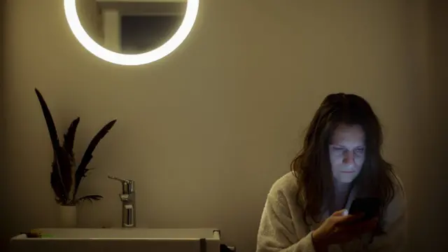 Una mujer revisa su teléfono dentro de un oscuro cuarto de baño