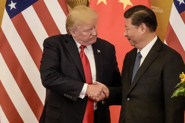 Trump apertando a mão de Xi Jinping com as bandeiras de seus respectivos países ao fundo