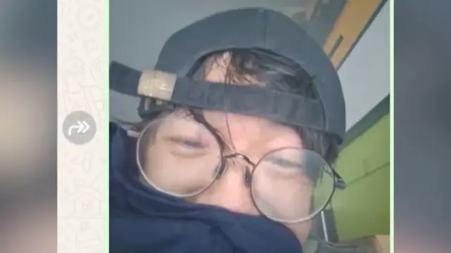 Вільям Лі у синьому одязі, він закриває рот і ніздрі. На ньому чорна бейсболка, одягнена задом наперед. Його окуляри запітнілі. Видно стіни та стелю його квартири позаду нього.