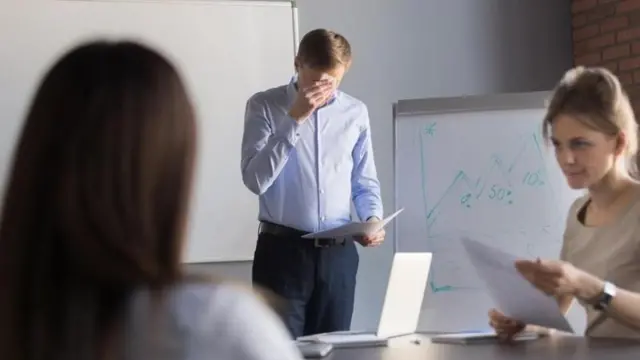 رجل يقف أمام زملائه في اجتماع عمل ويضع يده على وجهه، ربما لأنه يشعر بالتوتر أو الخجل.