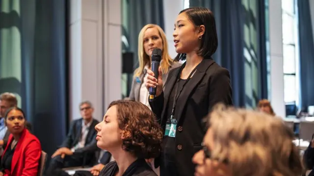 Une femme pose une question debout et tient un micro alors que d'autres personnes sont assises et qu'une femme se retourne pour la regarder.
