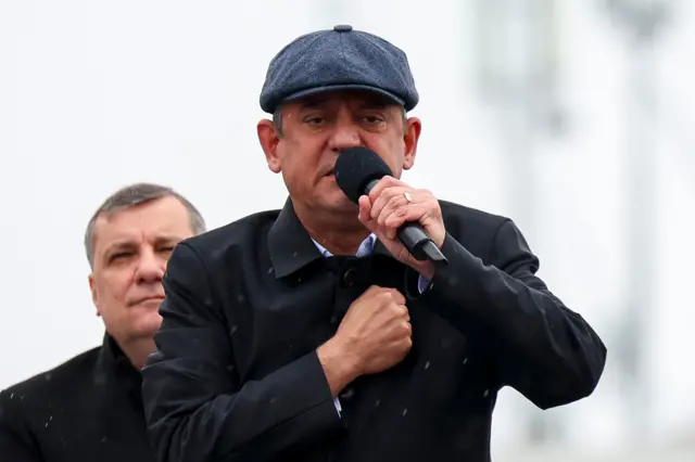 Özgür Özel kasket taktığı Çanakkale mitinginde 28 Mart'ta mikrofona konuşma yapıyor. Özel'in arkasından bir kişi görünüyıor