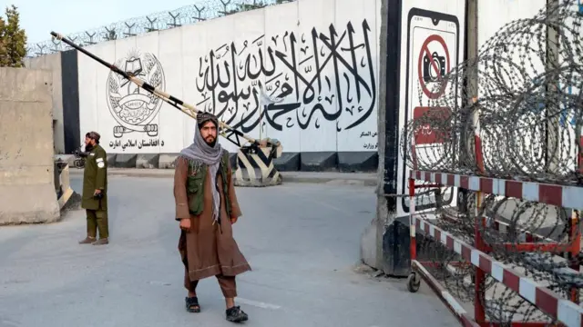 د طالبانو دوه ساتونکي برېښي چې د امر بالمعروف وزارت په مخه کې ولاړ دي او څېرمه د پاټک ستنه پورته شوې ده. 