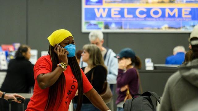 Passageira com máscara fala ao celular em aeroporto americano, com tela atrás exibindo a palavra 'Welcome'