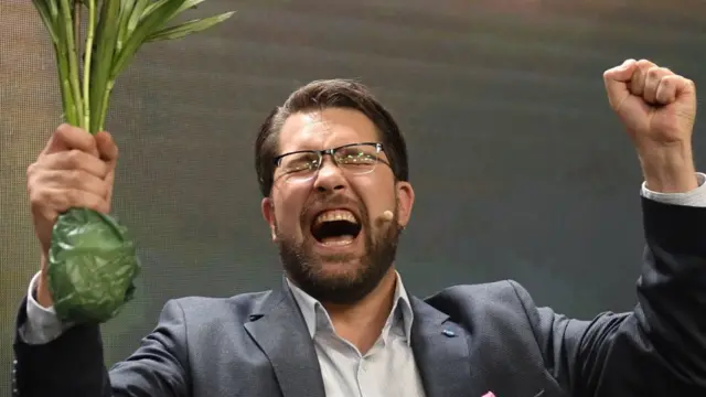 Foitografiaxbetinghomem branco com óculos, barba, cabelo castanho. Ele segura uma planta na mão direita e ergue o punho da esquerda, comemorando a vitória