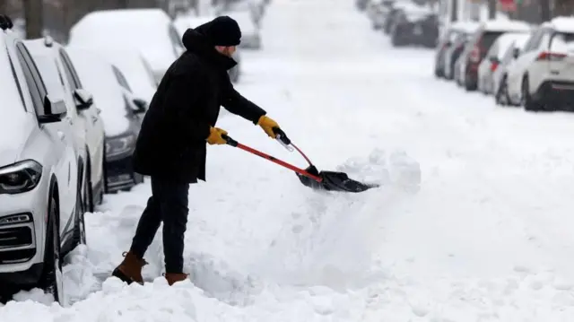 A man rugged up in heavy winter clothing uses a tool to clear snow packed into a shovel he is using to clear part of a road near a line of parked cars, all of which have snow over their roofs and windscreens.