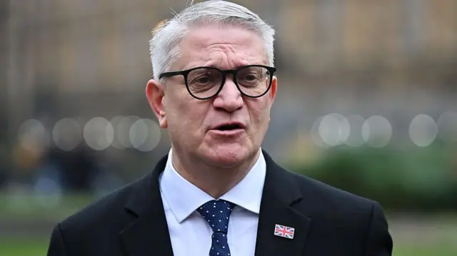 En la foto se ve a Andrew Rosindell con gafas, traje oscuro y corbata, hablando al aire libre, con árboles y barandillas borrosos en el fondo.