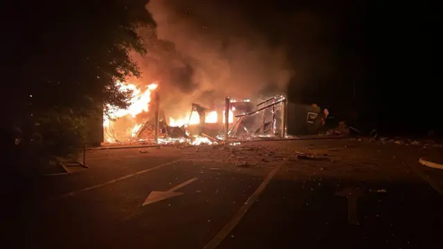 Уночі на дорозі палає зруйнована будівля або споруда. Яскраве полум'я охопило конструкцію, над нею піднімаються густі клуби диму. На асфальті перед пожежою видно уламки та дорожню розмітку зі стрілкою, що вказує напрямок руху.