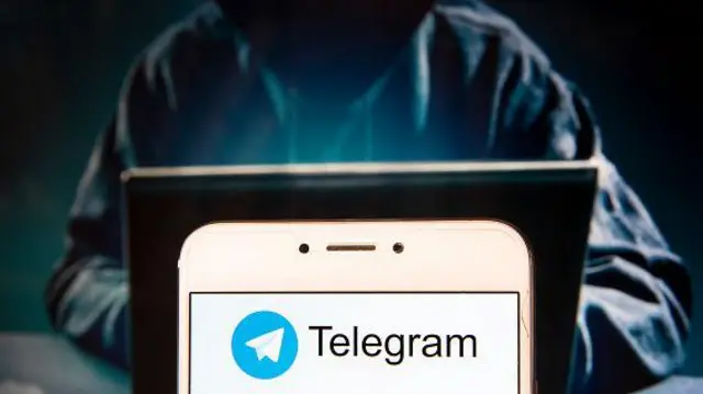 Колаж, на якому зображена людина у темному одязі, в капюшоні, який сидить за ноутбуком. На першому плані яскравий смартфон із логотипом телеграма. 