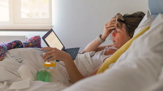 Uma pessoa deitada em uma cama, com um computador no colo, com os olhos fechados e uma toalha na testa. Ela segura um copo com um líquido laranja e ao redor dela há lenços de papel e comprimidos. 