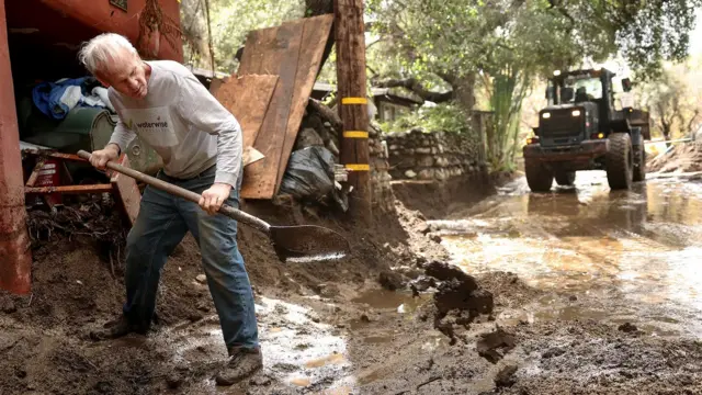 Homem com uma pá limpa a lama após deslizamento de terra na Califórnia, em fevereiro de 2025, com veículo de serviço ao fundo