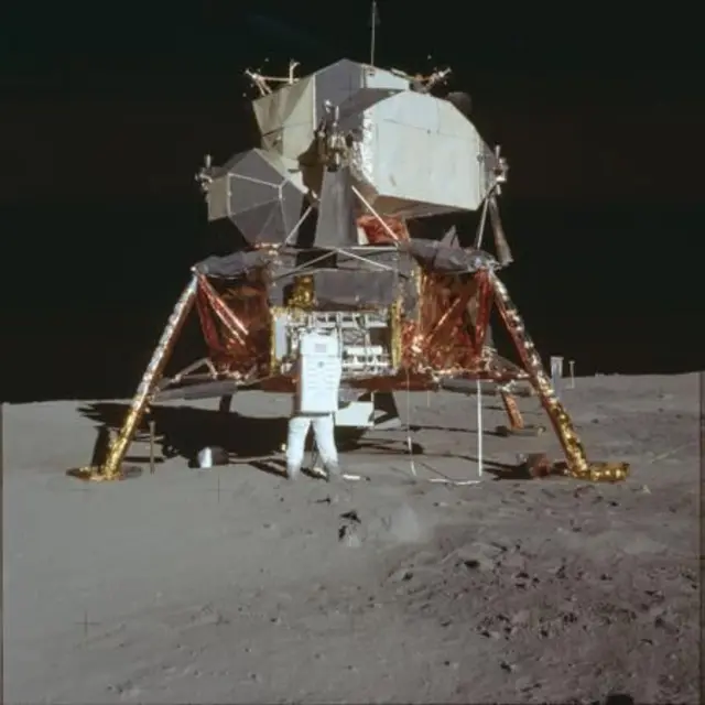 The lunar lander stands squat and insect‑like on spindly golden legs, its foil‑wrapped descent stage glowing copper against the ash‑grey dust. Above, the pale, faceted cabin looks almost improvised – a fragile aluminium refuge dropped into a pristine desert. The harsh sunlight carves inky black shadows beneath the lander and across the pitted regolith, where bootprints and equipment puncture an otherwise untouched plain. 