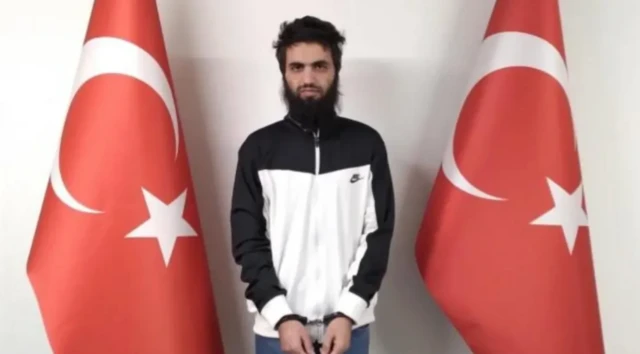 Elleri önden kelepçeli sakallı bir adam. İki yanında Türk bayrakları var. 