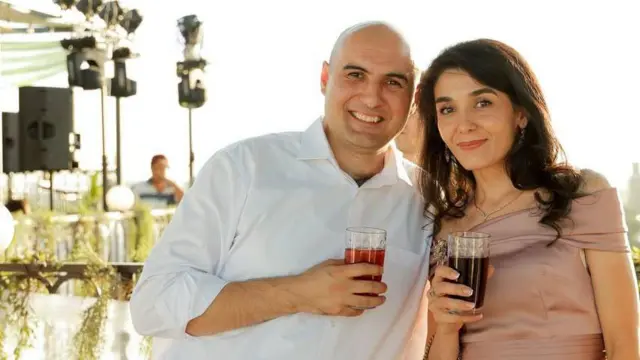 
Zafar e Dilya estão na foto tomando um drinque juntos. Ele veste uma camisa branca. Dilya usa uma blusa de seda rosa claro com decote ombro a ombro. Ela tem longos cabelos castanhos.