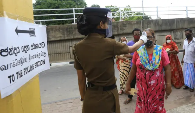 A Sri Lankan female police officer wearing a face mask conducts temperature checks as Sri Lankan voters wearing face masks stand in line during a mock election ahead of the parliamentary election in Colombo, Sri Lanka, on June 20, 2020.