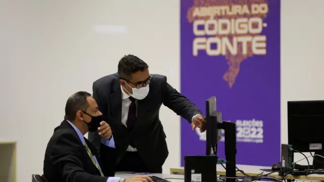 Dois homens olhando para computador, com painel atrás escrito 'Abertura do código-fonte'