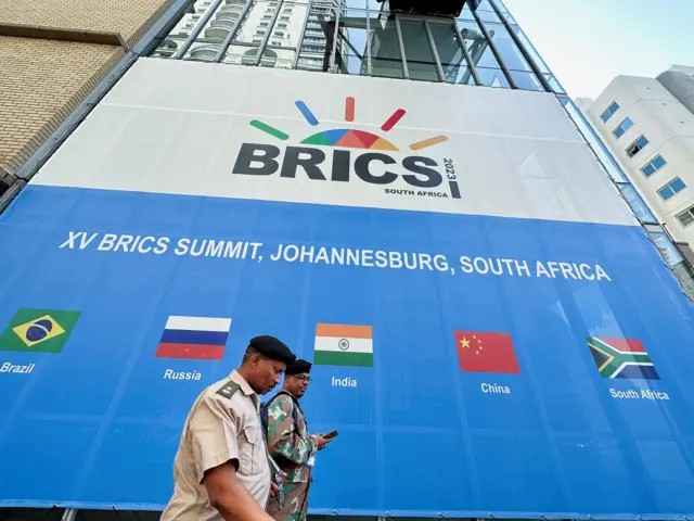 Dois homens passambwin o que significafrente a painelbwin o que significaevento dos BRICS que mostra cinco bandeiras dos membros