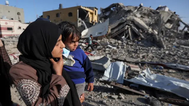 A Palestinian woman carries a child while they walk next to destroyed houses in Khan Younis, Gaza