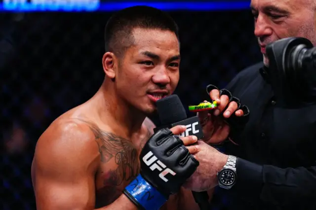 မြန်မာနိုင်ငံ အလံအရောင်တွေပါတဲ့ အံကပ်ကို လက်ကကိုင်ပြီး မိုက်နဲ့စကားပြောနေတဲ့ ဂျော့ရှုဝါဖန်