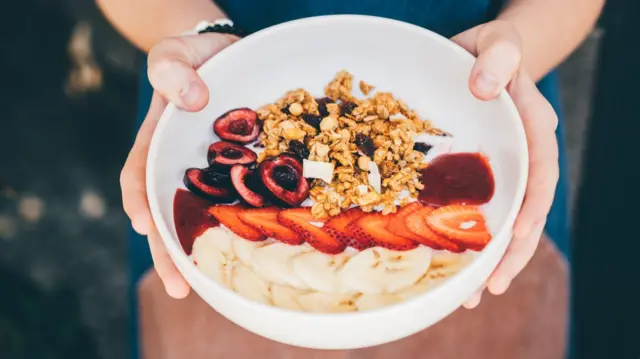 
په دې عکس کې د یوه کس لاسونه ښکاري چې سپین رنګه کاسه یې نیولې ده او ځمکني توتان، کیله او یو څه نوره میوه په کې ده. 