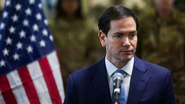 مارکو روبیو، وزیر خارجه آمریکا با کت و کراوات آبی در کنار پرچم آمریکا جلوی سربازان اسرائیلی با لباس نظامی