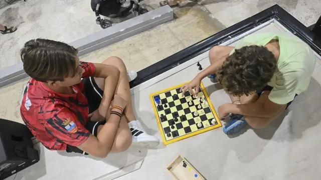Dos niños vistos desde arriba, van vestidos con ropa deportiva de verano y zapatillas deportivas. Están sentados uno frente a otro, con las piernas cruzadas. Tienen delante de ellos un tablero de ajedrez y están jugando una partida. 