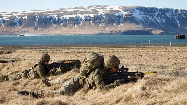 Dois soldados americanos estão deitados, apontando suas armas em um campo. A que está atrás parece ser uma mulher. Ao fundo, vê-se montanhas nevadas.