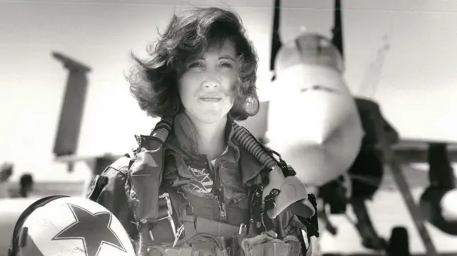 Donanma taktik uçaklarını kullanan ilk kadınlardan biri olan Teğmen Tammie Jo Shults, bir F/A-18'in önünde poz veriyor. Pilot tulumu giymiş ve kaskını sağ elinde tutuyor.