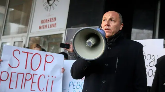 Андрій Парубій в 2020 році - в чорному пальті з рупором на фоні плакату, на якому написано "STOP репресії"