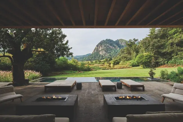 Terraza contemporánea cubierta con camas lounge y fogones, abierta a un paisaje verde con montañas al fondo.