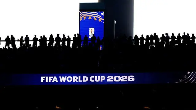 A dark image of a stand at the FIFA World Cup against a light background with a blue illumination of a World Cup logo on a pillar behind the crowd. 