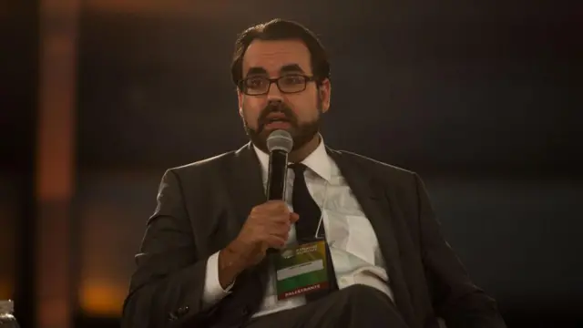 Christopher Garman sentado e falando ao microfone durante um evento.