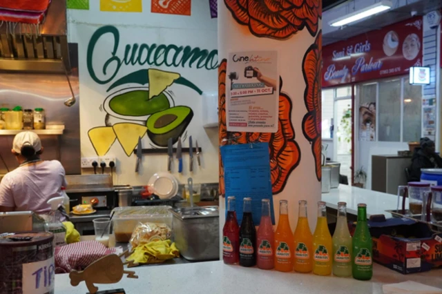 Se ve un mostrador del restaurante. y sobre este una hilera de botellas con soda. en la pared del fondo hay una ilustración de un aguacate con tortillas y la palabra "guacamole". Se a una empleada de espaldas cocinando. 